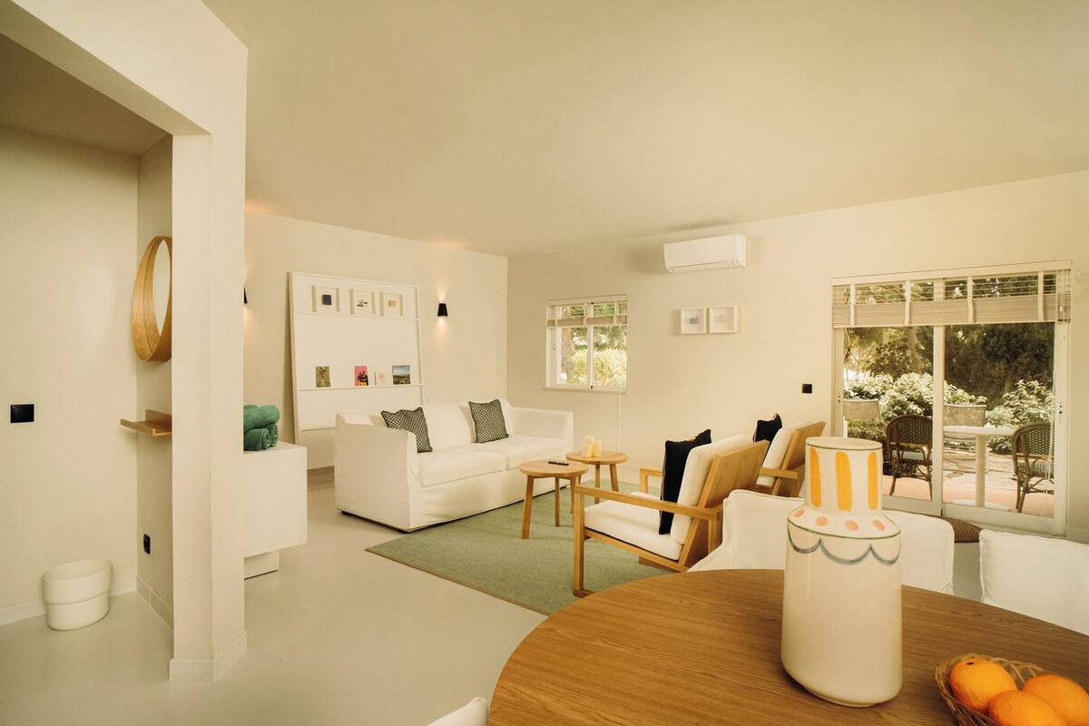 A modern living room with a white sofa, a wooden dining table, and a view of the outdoors through large windows.