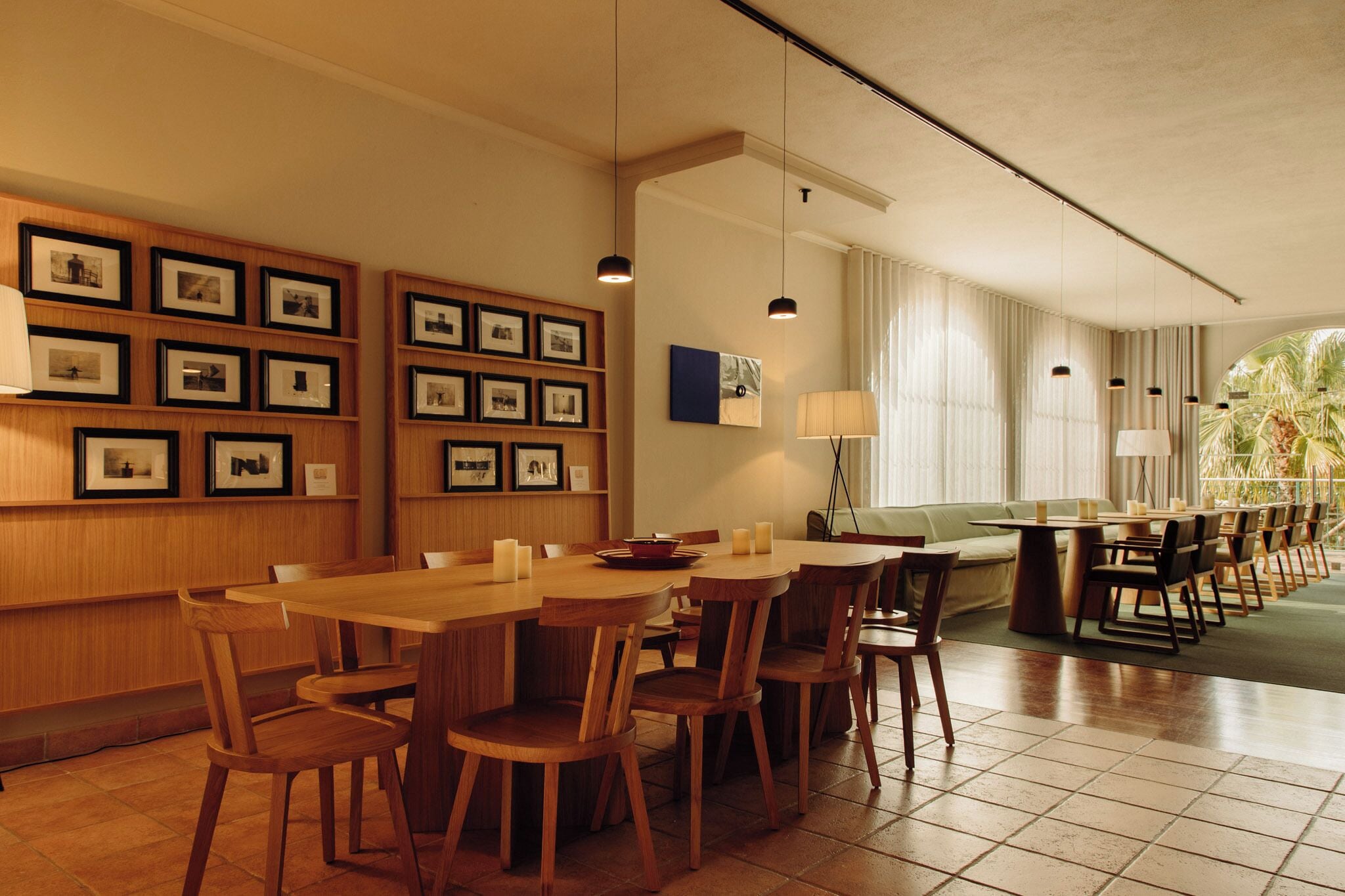 Un restaurant avec des tables et des chaises en bois, des tableaux encadrés aux murs et une vue sur les palmiers par les fenêtres.