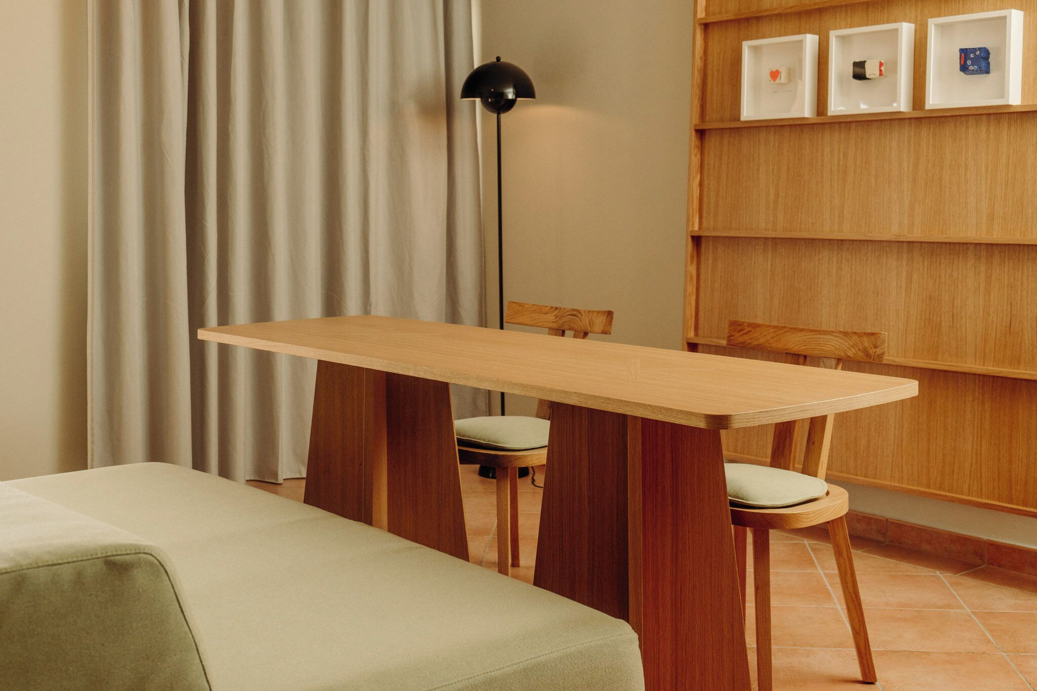 A room with a wooden table, two chairs, a floor lamp, and a shelf with framed items.