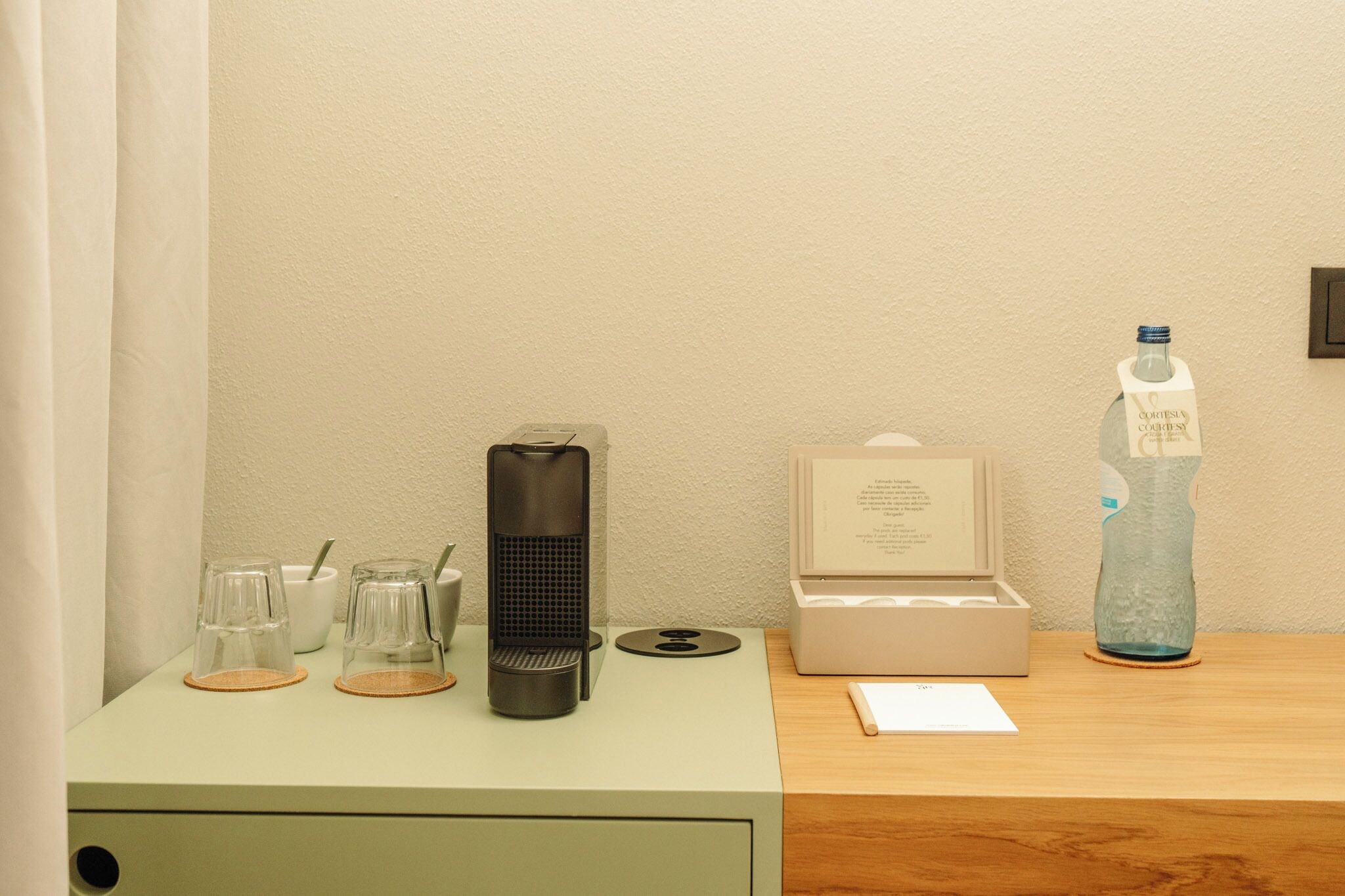 A hotel room with a desk featuring a coffee maker, a water bottle, a box of tissues, and a small table with a cup and saucer.