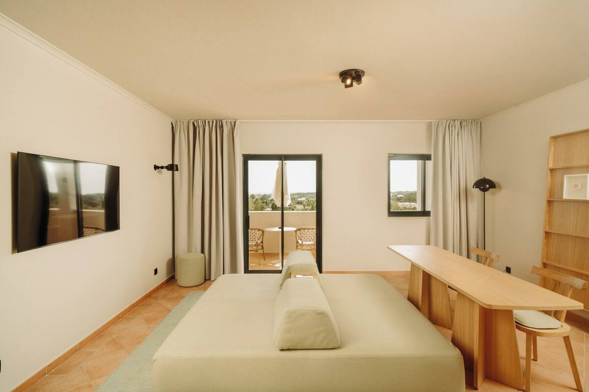 A modern hotel room with a bed, a dining table, chairs, a TV, and a balcony with a view.