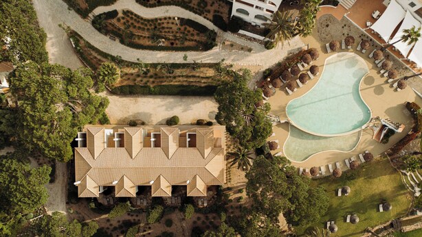 Vista aérea de un complejo turístico con una piscina central, varias zonas de descanso con sombrillas y un jardín ajardinado.