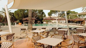 Un área junto a la piscina con sillones de descanso y mesas, una piscina y palmeras.