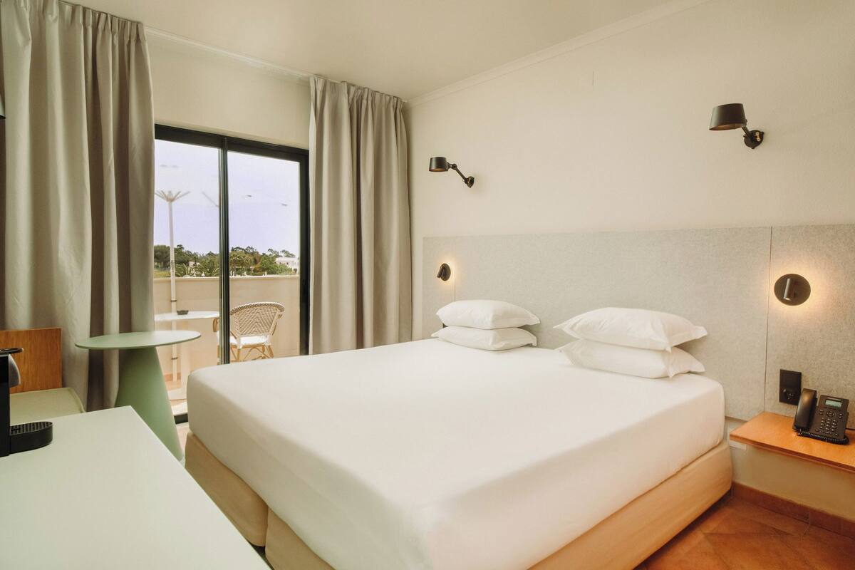 A hotel room with a bed, bedside table, lamp, and a view of the outdoors.