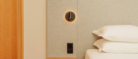 Une chambre avec une table de chevet en bois, une lampe fixée au mur et un lit recouvert de linge de lit blanc.