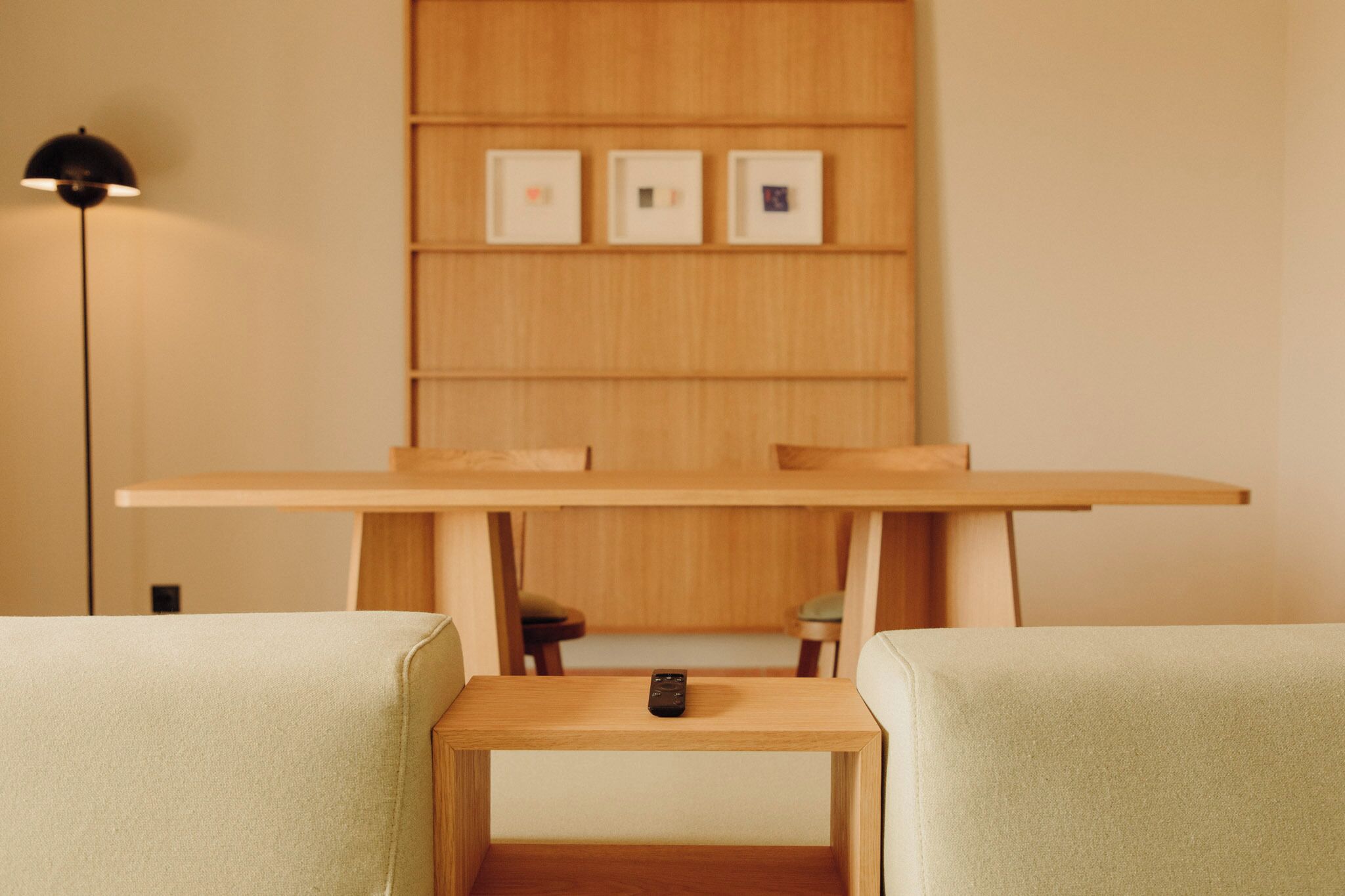Une pièce au décor minimaliste, avec une table en bois, deux chaises rembourrées, une télécommande posée sur la table et des tableaux encadrés accrochés au mur.