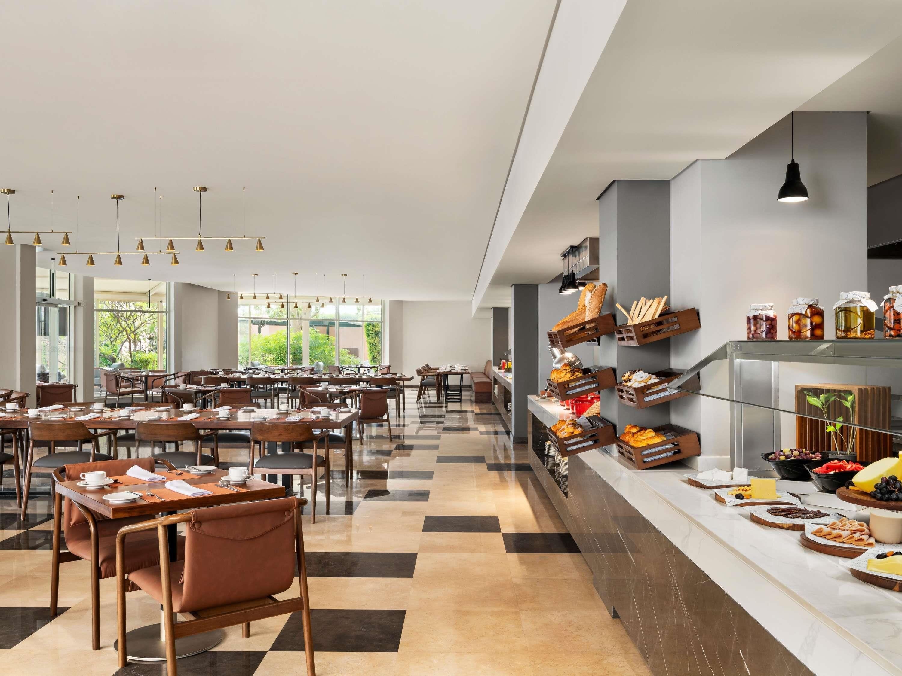 A spacious dining area with multiple tables and chairs, a buffet counter with various food items, and large windows providing natural light.