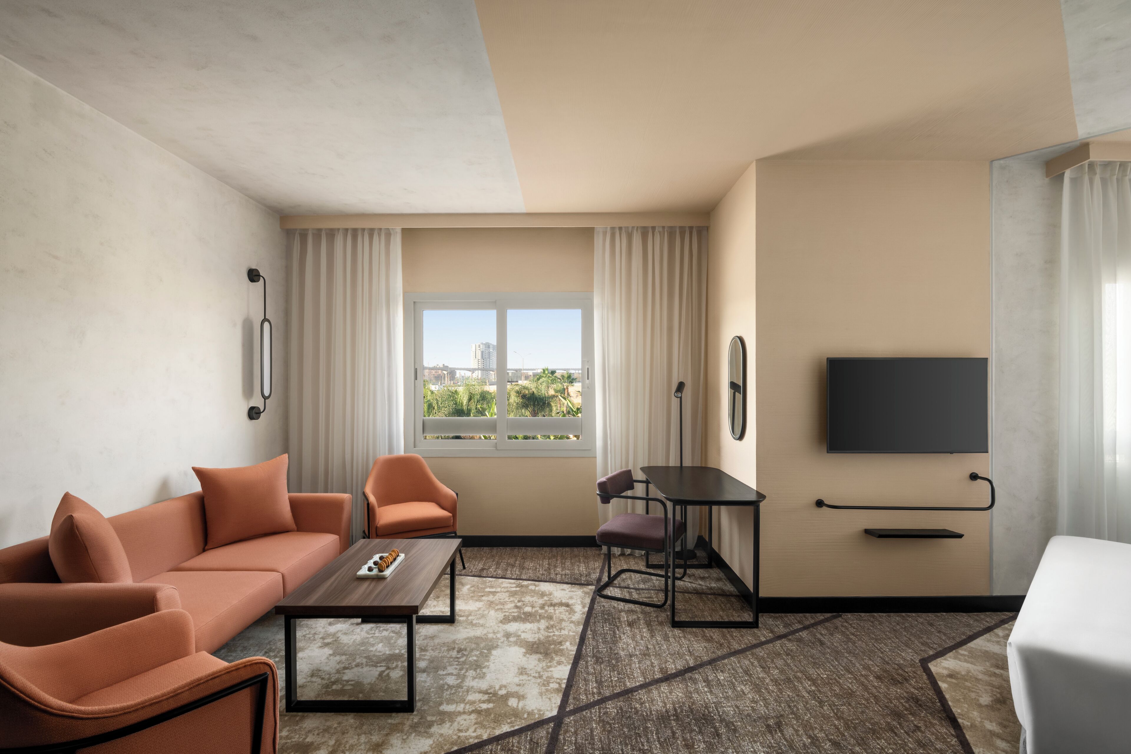 A hotel room with a sofa, armchairs, a dining table, a TV, and a large window with a city view.