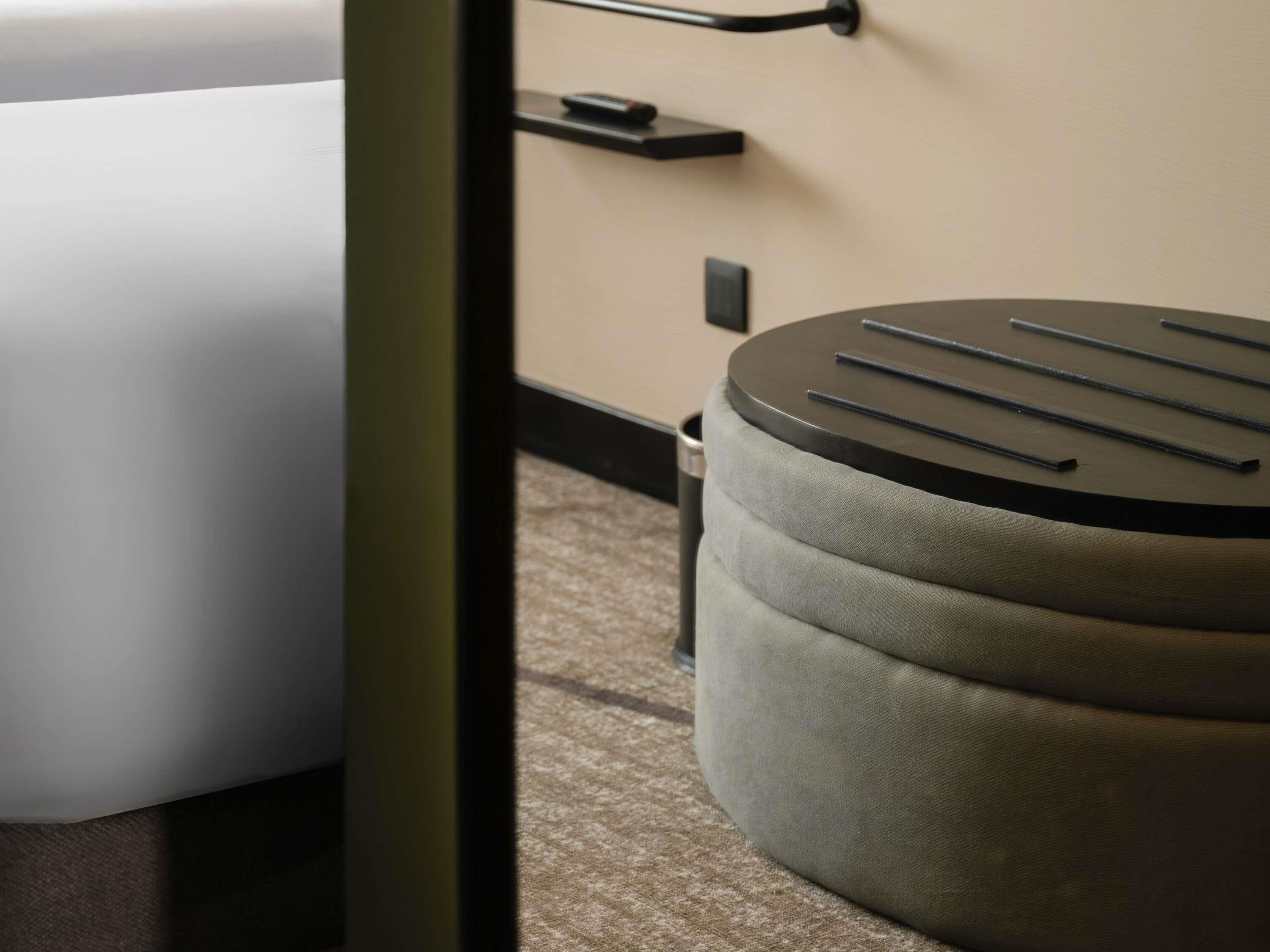 A hotel room with a bed, a round ottoman with a black top, a wall-mounted shelf, and a door with a handle.
