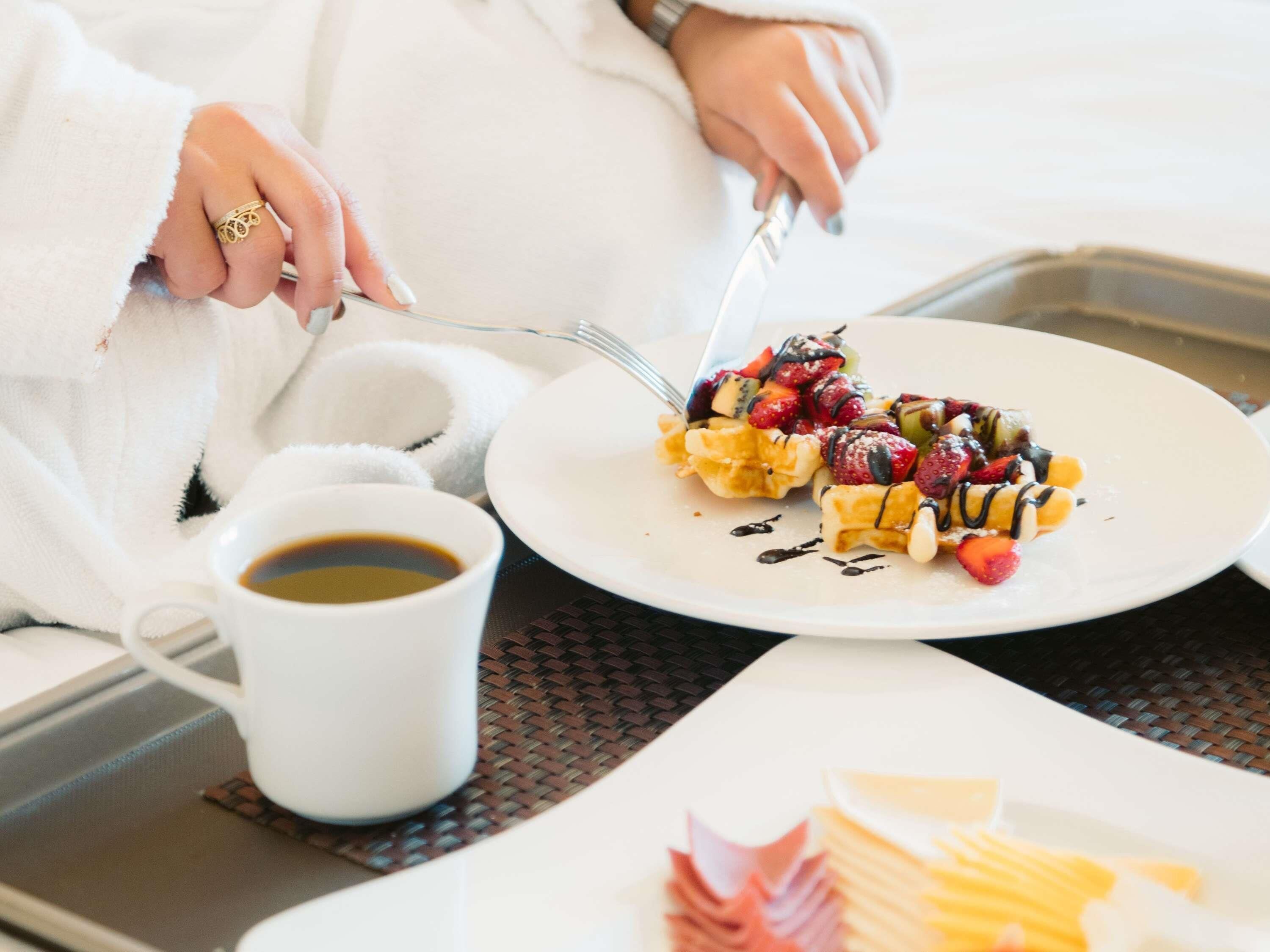 Une personne vêtue d’une robe blanche est en train de prendre son petit-déjeuner : son assiette contient des gaufres, des fruits et une sauce au chocolat. Une tasse de café se trouve à côté d’elle.