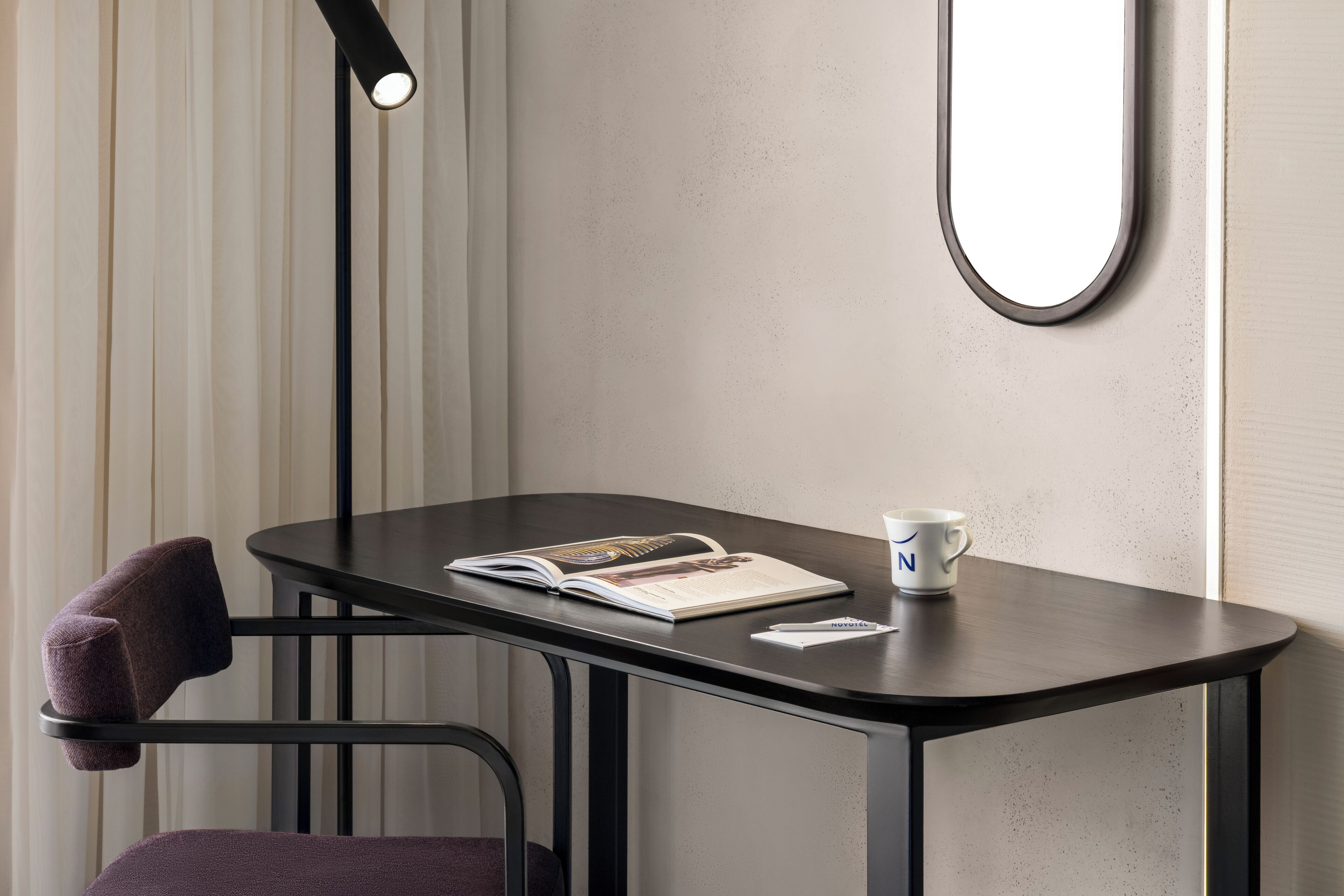 Une petite table en bois sombre, sur laquelle se trouvent un magazine, un stylo et une tasse. Une chaise avec un coussin est placée devant la table.