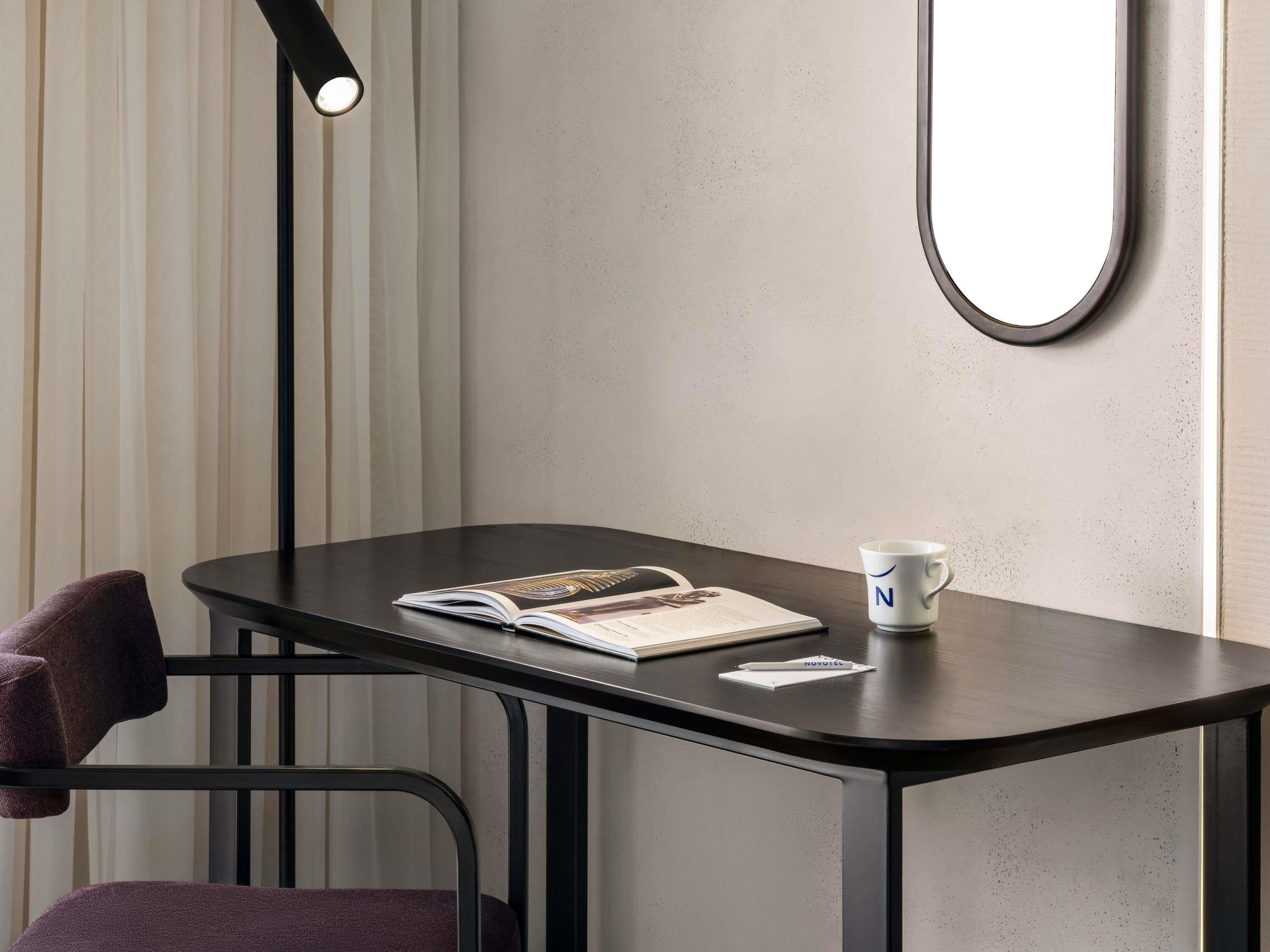 Une pièce d’angle avec une table en bois foncé, un magazine, une tasse blanche ornée d’une lettre, une chaise noire et un lampadaire.