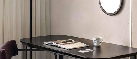 Une pièce d’angle avec une table en bois foncé, un magazine, une tasse blanche ornée d’une lettre, une chaise noire et un lampadaire.