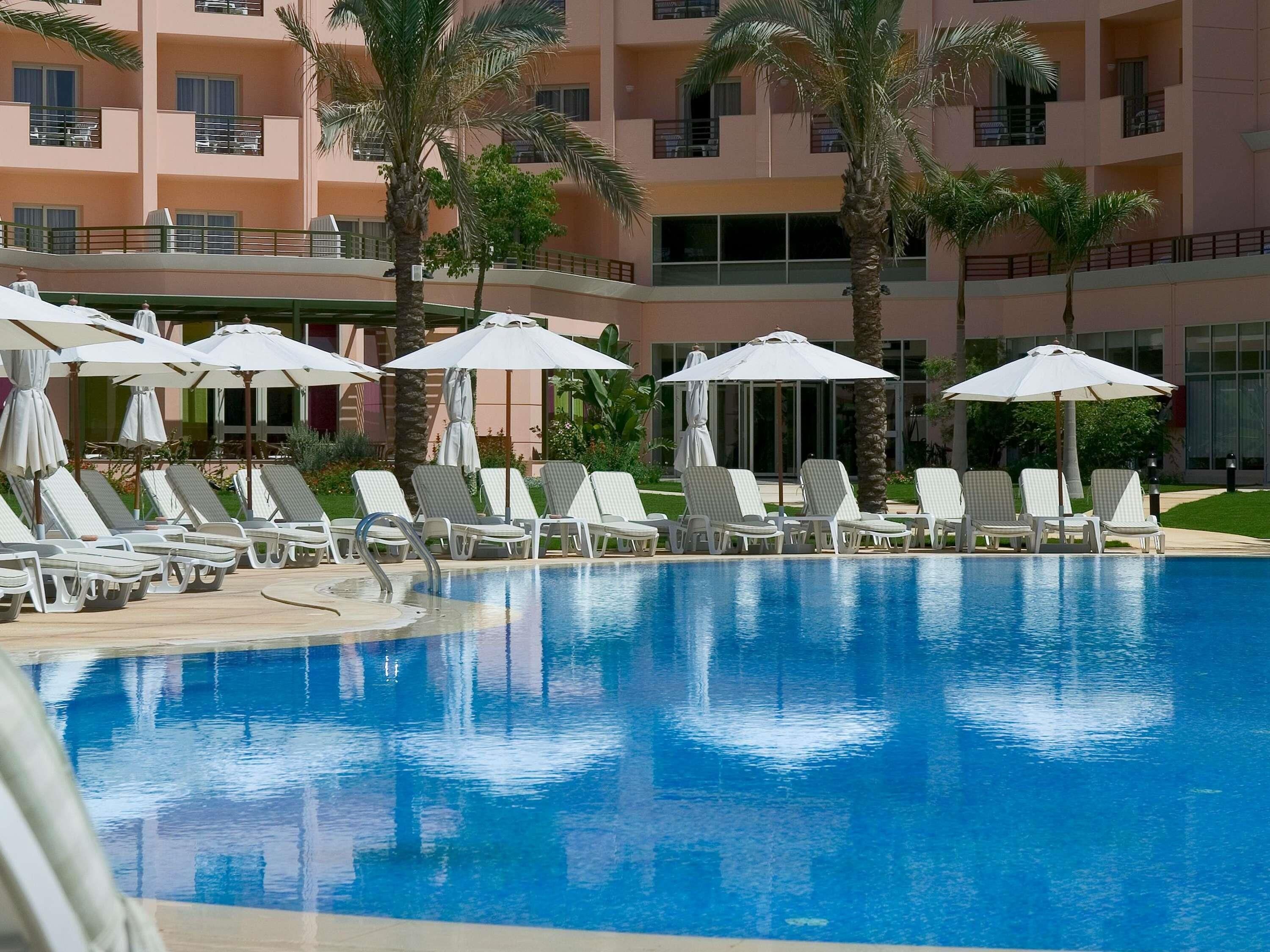 A swimming pool with lounge chairs and umbrellas, surrounded by palm trees and a multi-story building.