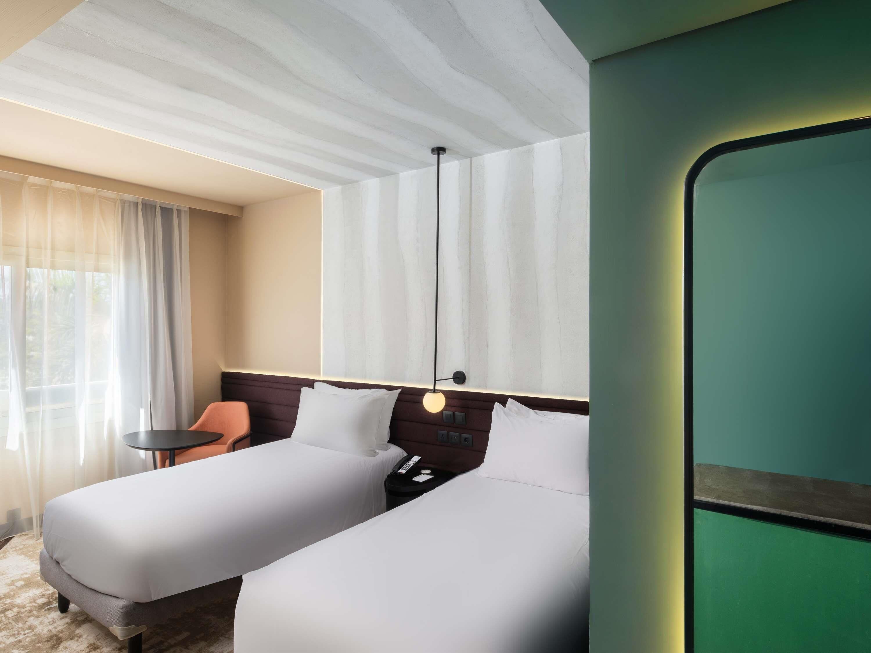 Habitación de hotel con dos camas, una mesita, una silla y una ventana con cortinas.