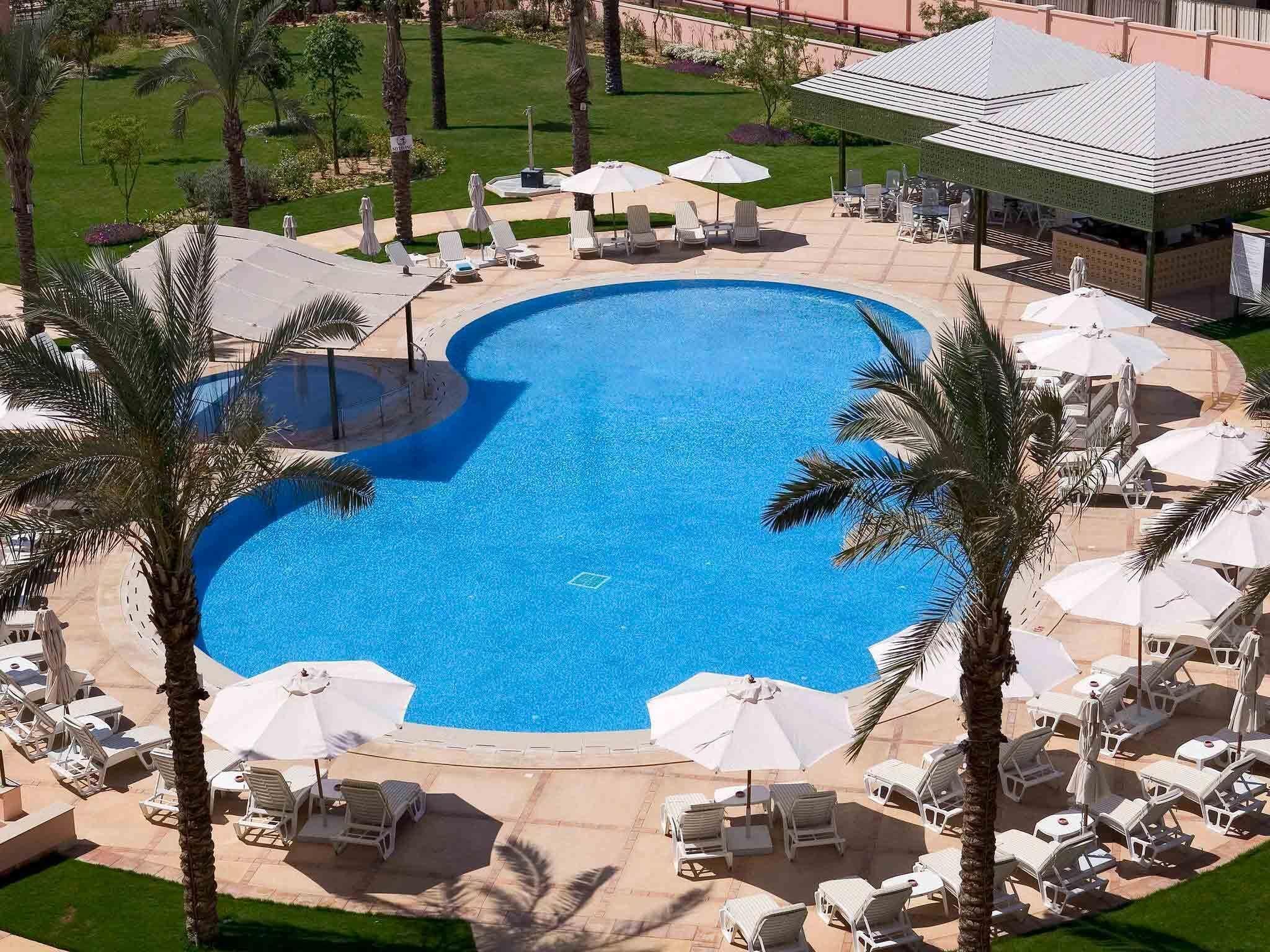 A large swimming pool surrounded by lounge chairs and umbrellas, with palm trees and a well-maintained lawn.