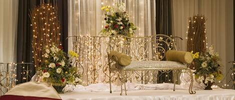 A banquet hall with round tables set for dining, a stage with a backdrop, and a decorative chandelier.