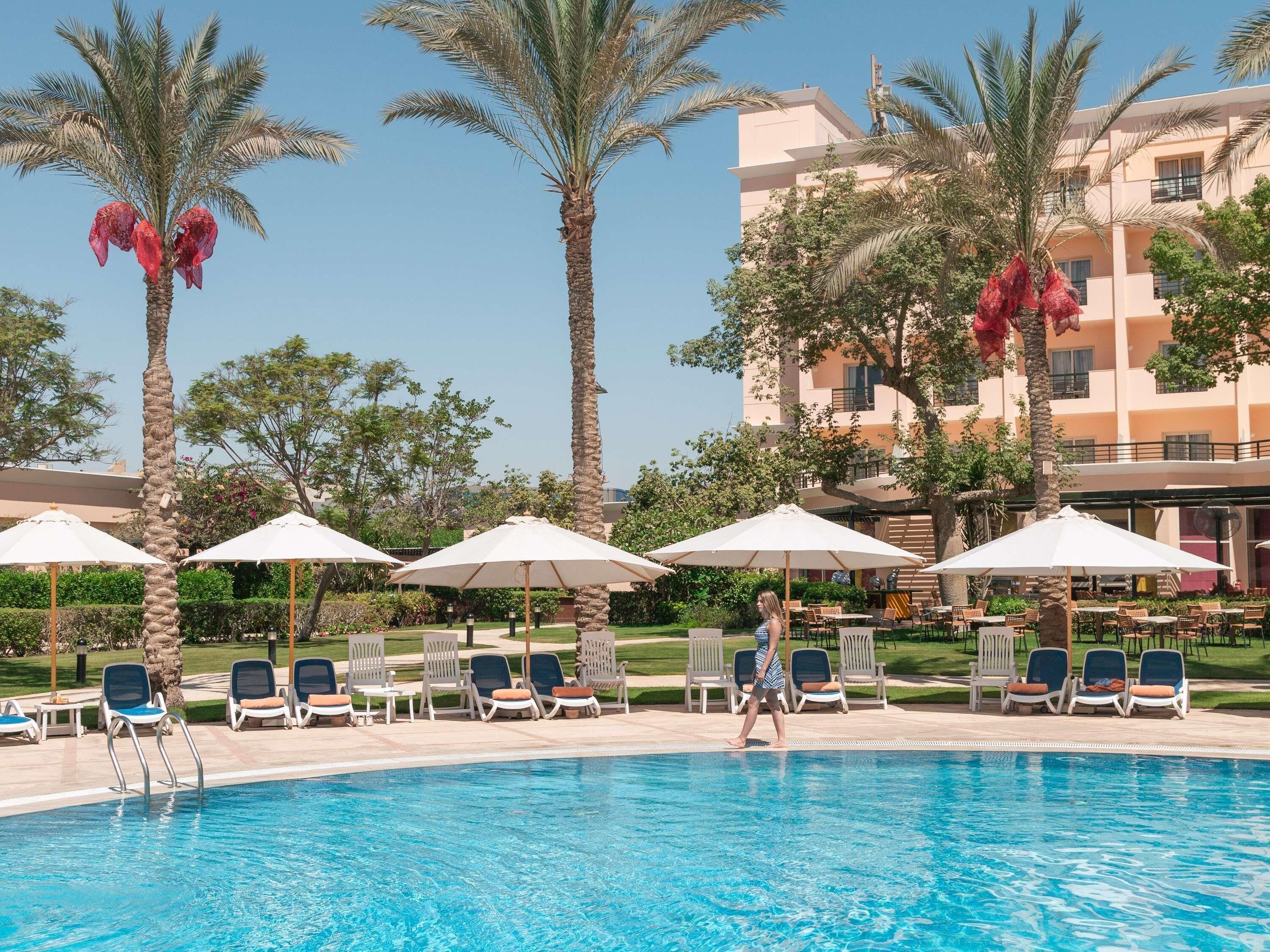 A poolside area with lounge chairs and umbrellas, surrounded by palm trees and a building in the background.