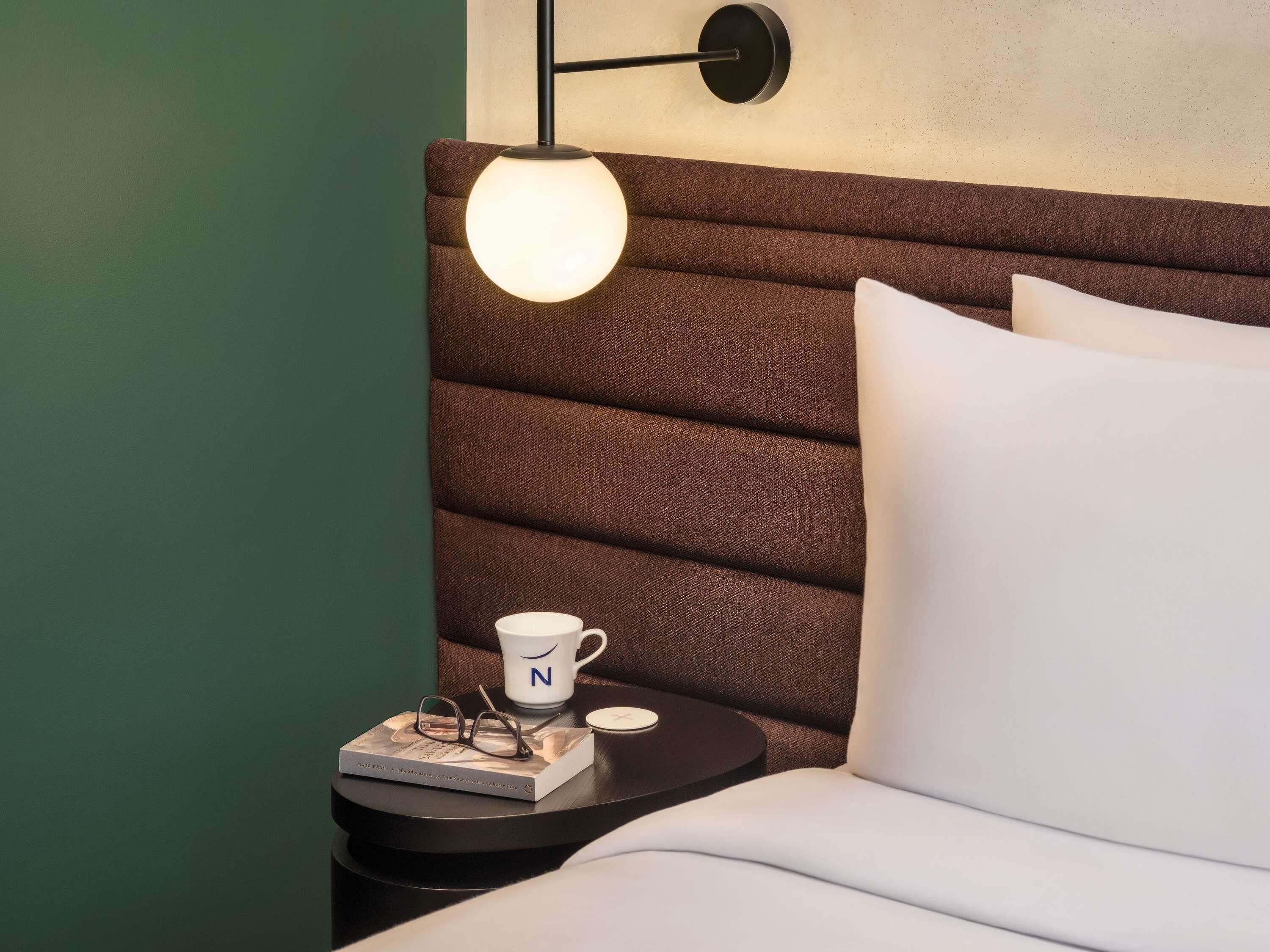 A neatly made bed with white pillows, a bedside table with a book, glasses, and a mug, and a wall-mounted lamp.