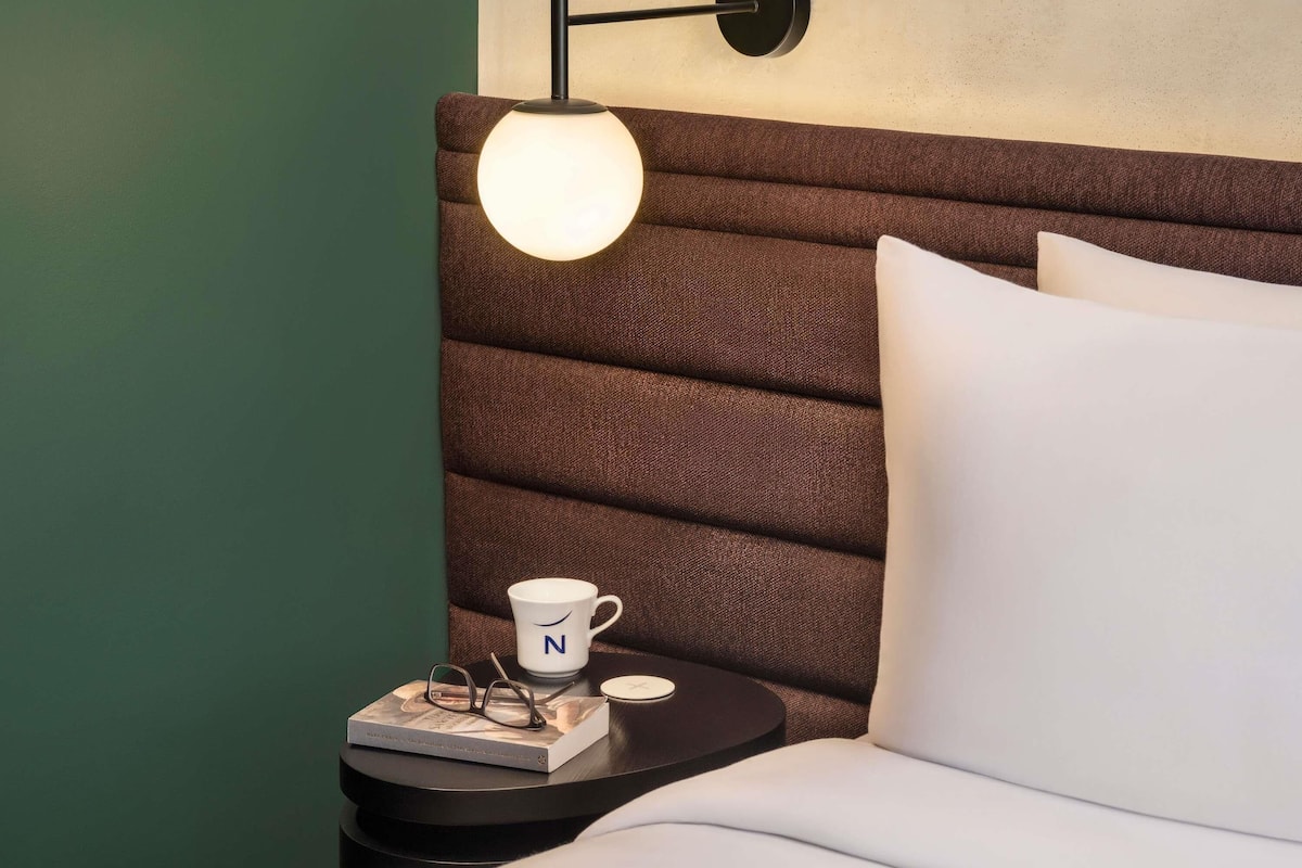 A neatly made bed with white pillows, a bedside table with a book, glasses, and a mug, and a wall-mounted lamp.
