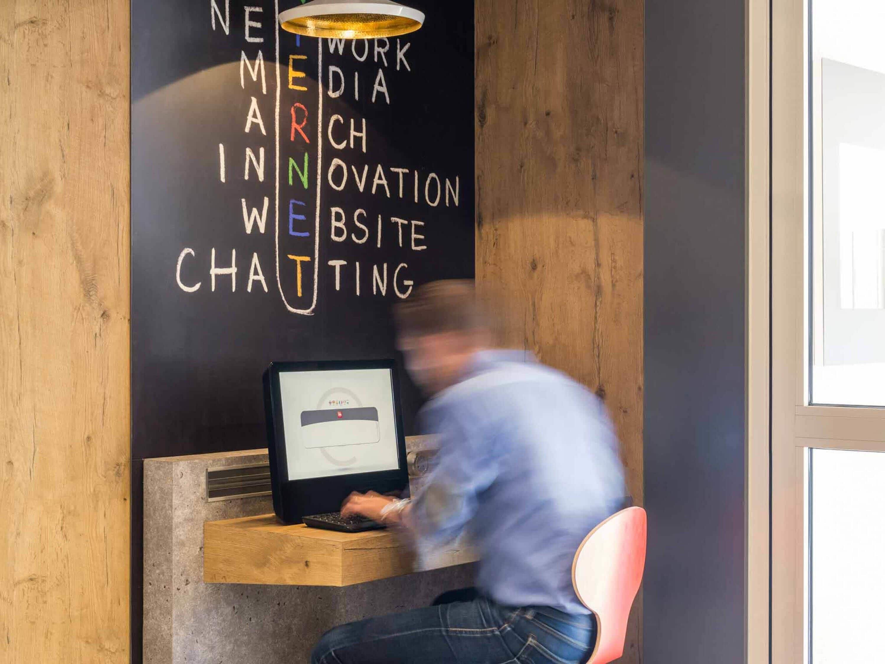 Una persona trabajando en un escritorio con una computadora, una pared de madera y una pizarra con palabras como 'WORK', 'MACHINE', 'OVATION', 'SITE' y 'CHATING' escritas en ella.