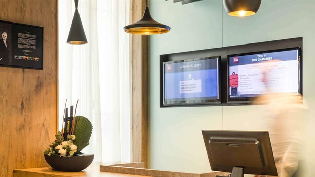 A hotel reception desk with two digital screens displaying room information, a black pendant light, and a wooden reception counter. - ibis Paris Levallois Perret (Levallois-Perret)
