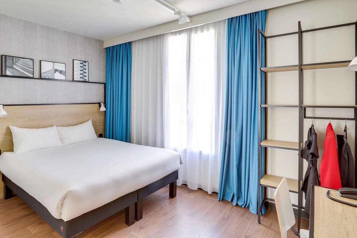 Habitación de hotel con cama, escritorio con silla, estante con ropa y ventana con cortinas.