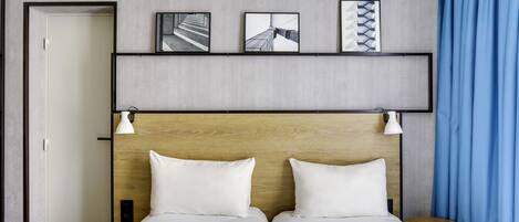 A hotel room with two beds, a wooden headboard, a small side table, and a wall-mounted shelf with framed pictures.