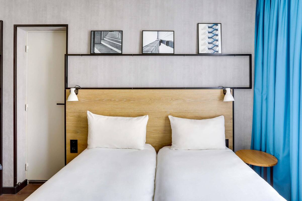Habitación de hotel con dos camas, cabecero de madera, estante empotrado con fotografías enmarcadas, cortina azul y una pequeña mesa redonda.