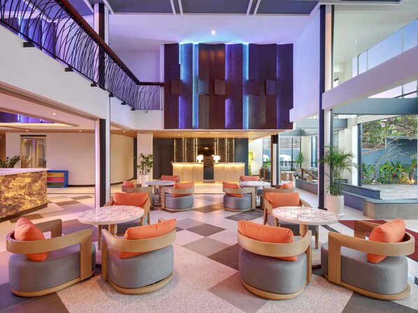Un vestíbulo moderno con mostrador de recepción, zona de estar con mesas redondas y sillones, y una escalera con un diseño de barandilla distintivo.