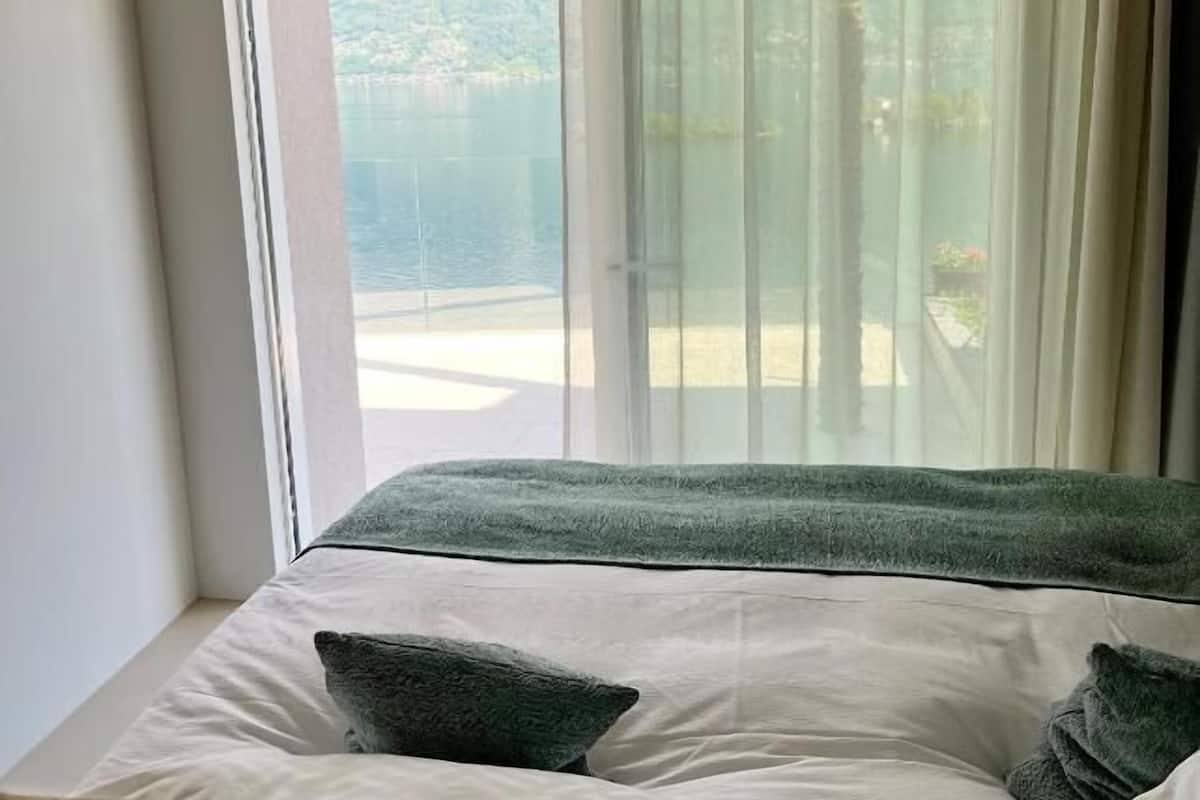 A bedroom with a large window, a bed with white linens, and a green throw blanket.