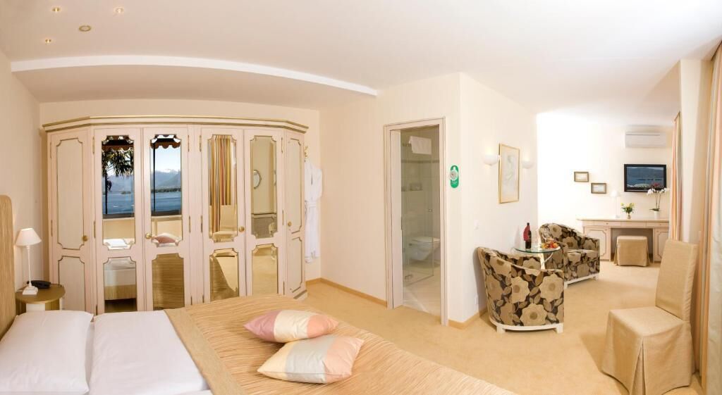 Habitación de hotel con cama, zona de estar con sofá y sillones, escritorio con silla, televisor y vista al mar a través de una puerta.