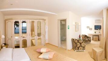 Habitación de hotel con cama, zona de estar con sofá y sillones, escritorio con silla, televisor y vista al mar a través de una puerta.