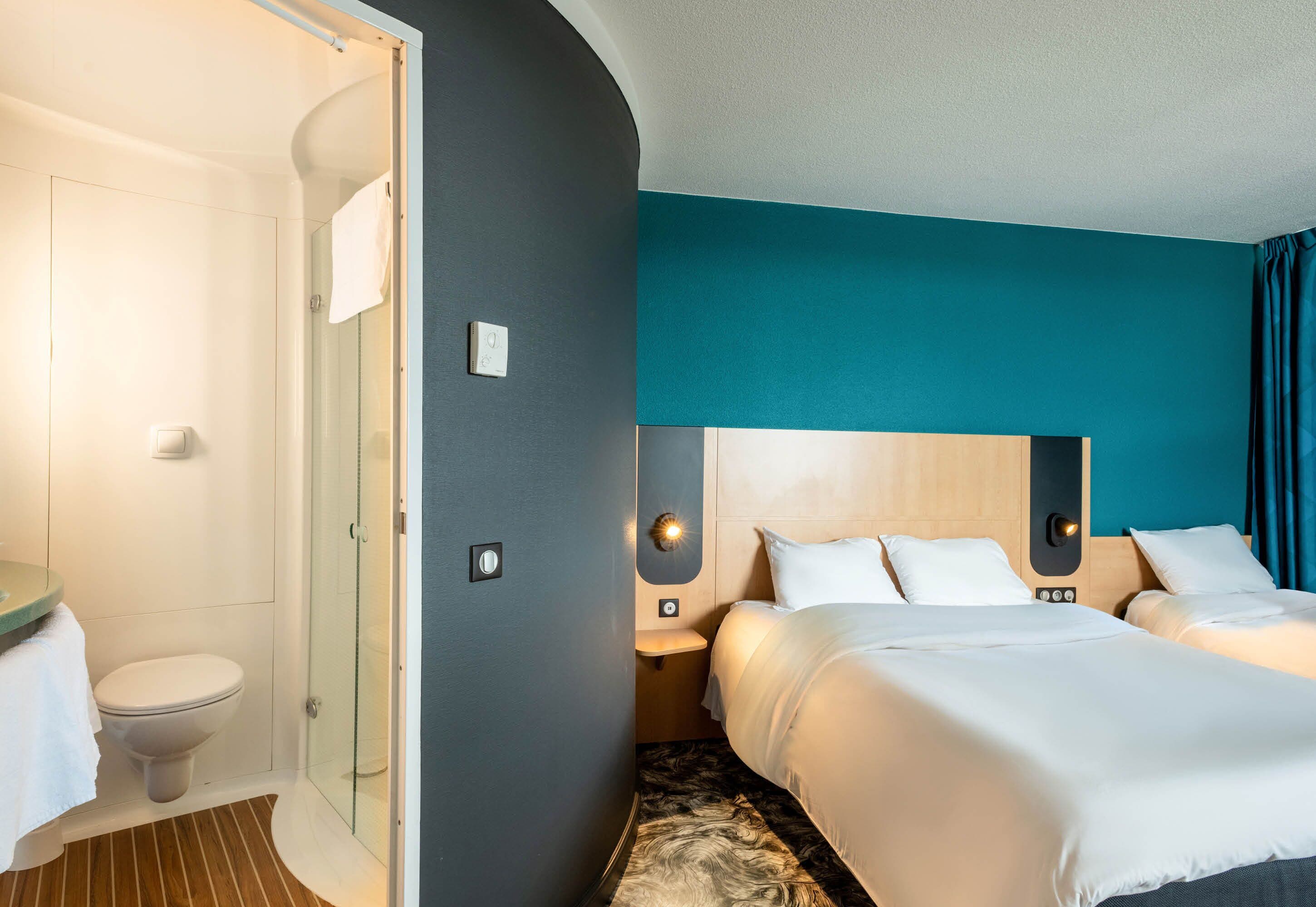 Une chambre d’hôtel équipée d’une douche, de toilettes, d’un lit avec une literie blanche et d’un mur d’accent de couleur turquoise.