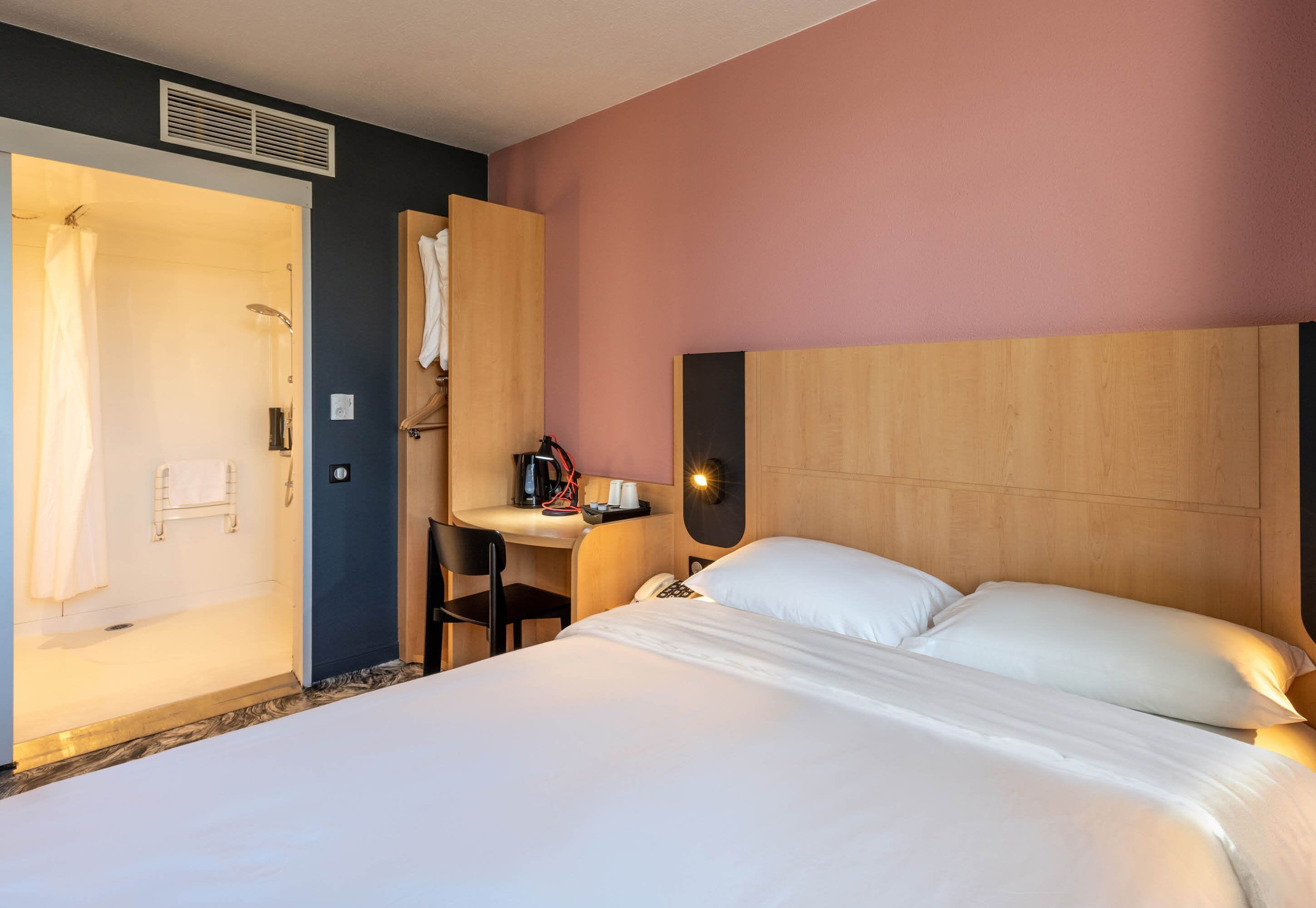A hotel room with a bed, a desk, a chair, a shower, and a towel rack.