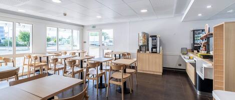 A spacious dining area with multiple tables and chairs, a kitchen area with appliances, and large windows providing natural light.
