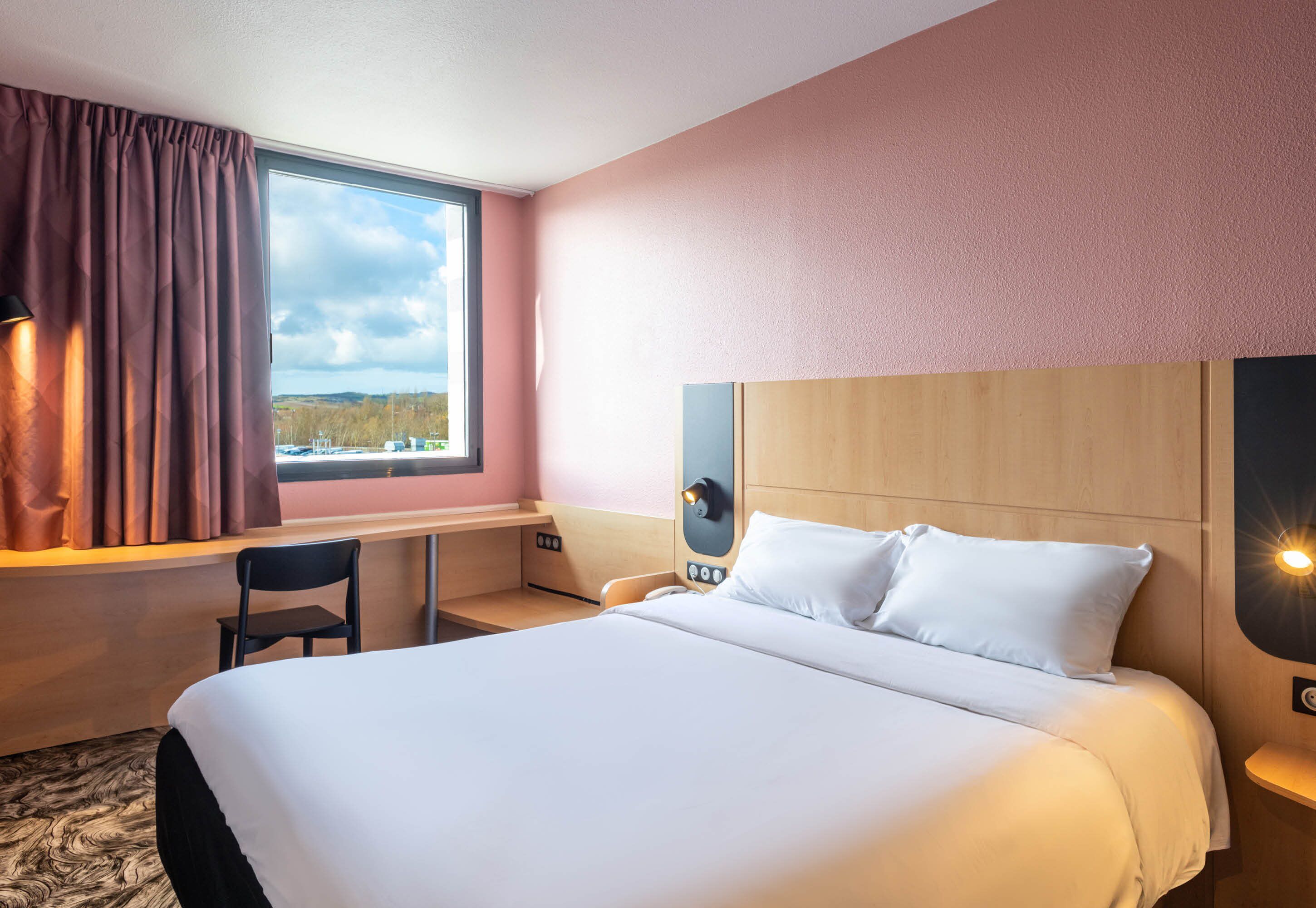 A hotel room with a large bed, a desk, a chair, a window with curtains, and a view of the outdoors.