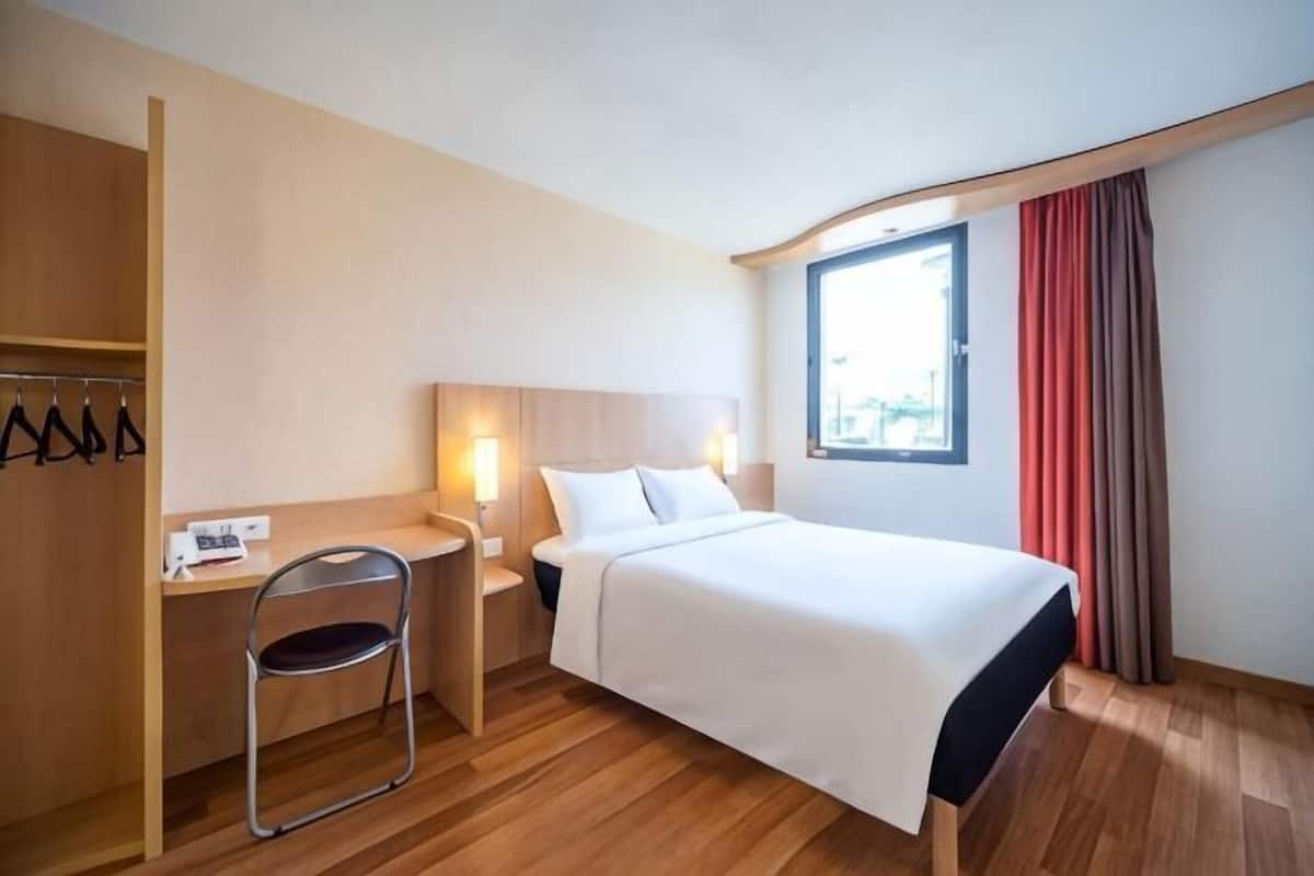 A hotel room with a bed, a desk with a chair, a nightstand, and a window with curtains.