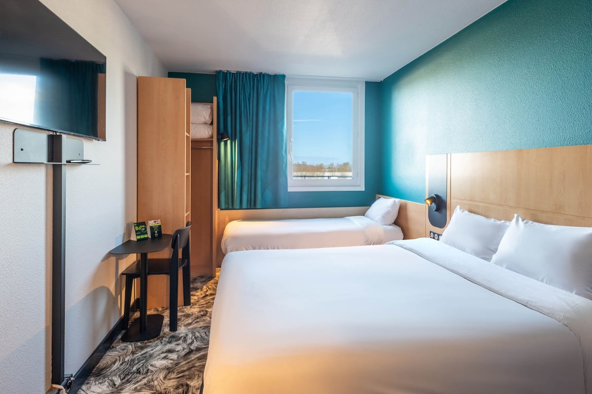 A hotel room with two beds, a desk with chairs, a TV, and a window with curtains.