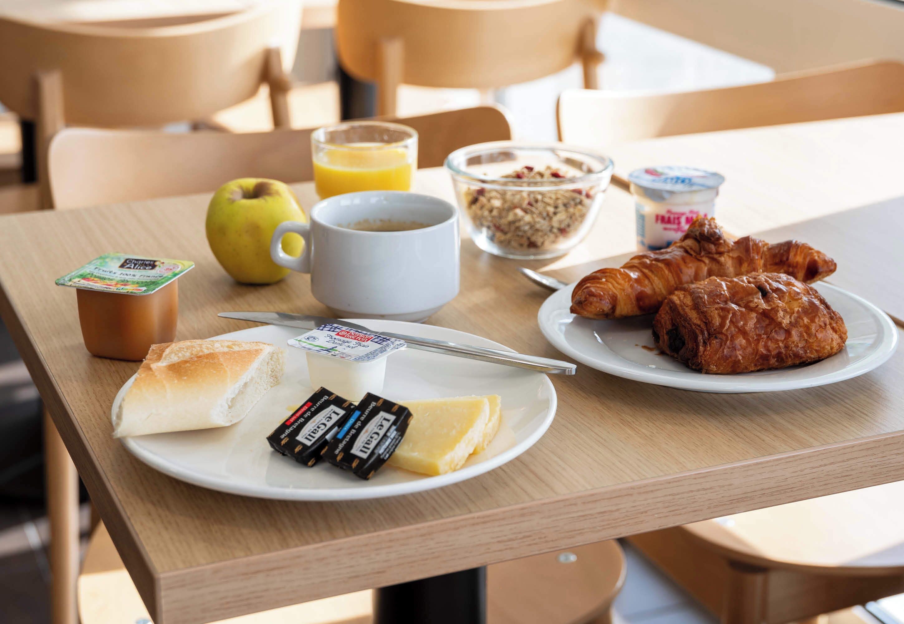 A breakfast table with croissants, cheese, butter, a bottle of juice, a bowl of cereal, and a cup of coffee.