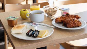 A breakfast table with croissants, cheese, butter, a bottle of juice, a bowl of cereal, and a cup of coffee.