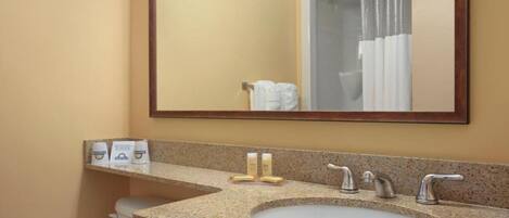 A bathroom with a granite countertop sink, two faucets, and a mirror above it. There are toiletries on the countertop and a shower curtain visible in the mirror reflection.