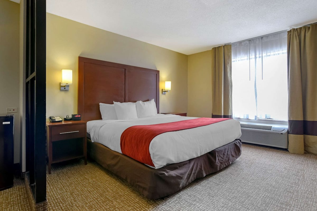 Une chambre d’hôtel avec un grand lit, deux tables de chevet, une petite table, une lampe fixée au mur, un appareil de climatisation et une fenêtre avec des rideaux.