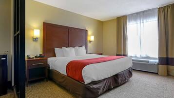 A hotel room with a large bed, two bedside tables, a nightstand, a wall-mounted lamp, an air conditioning unit, and a window with curtains.