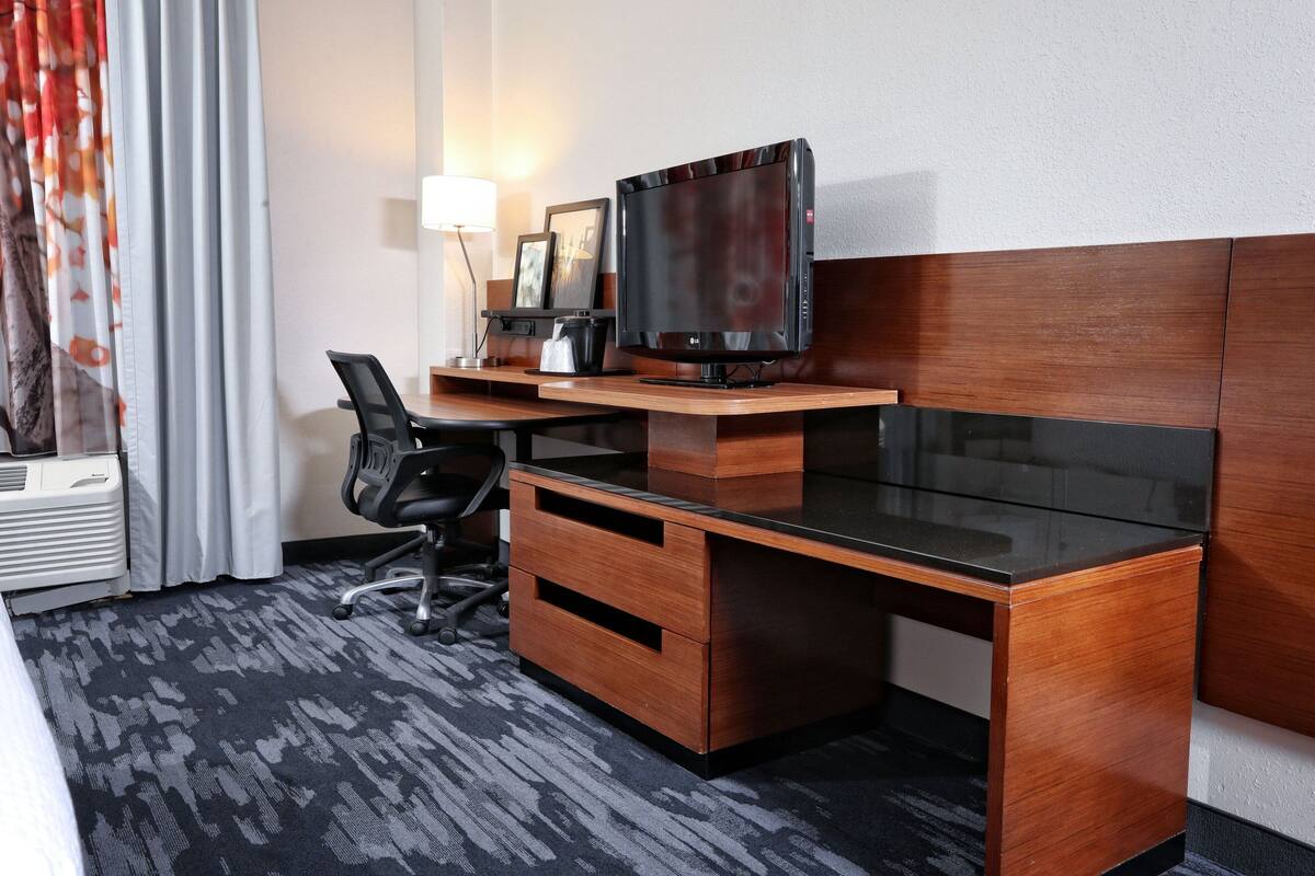 A hotel room with a wooden desk, a flat-screen TV, a chair, a lamp, and a bed with a patterned carpet.