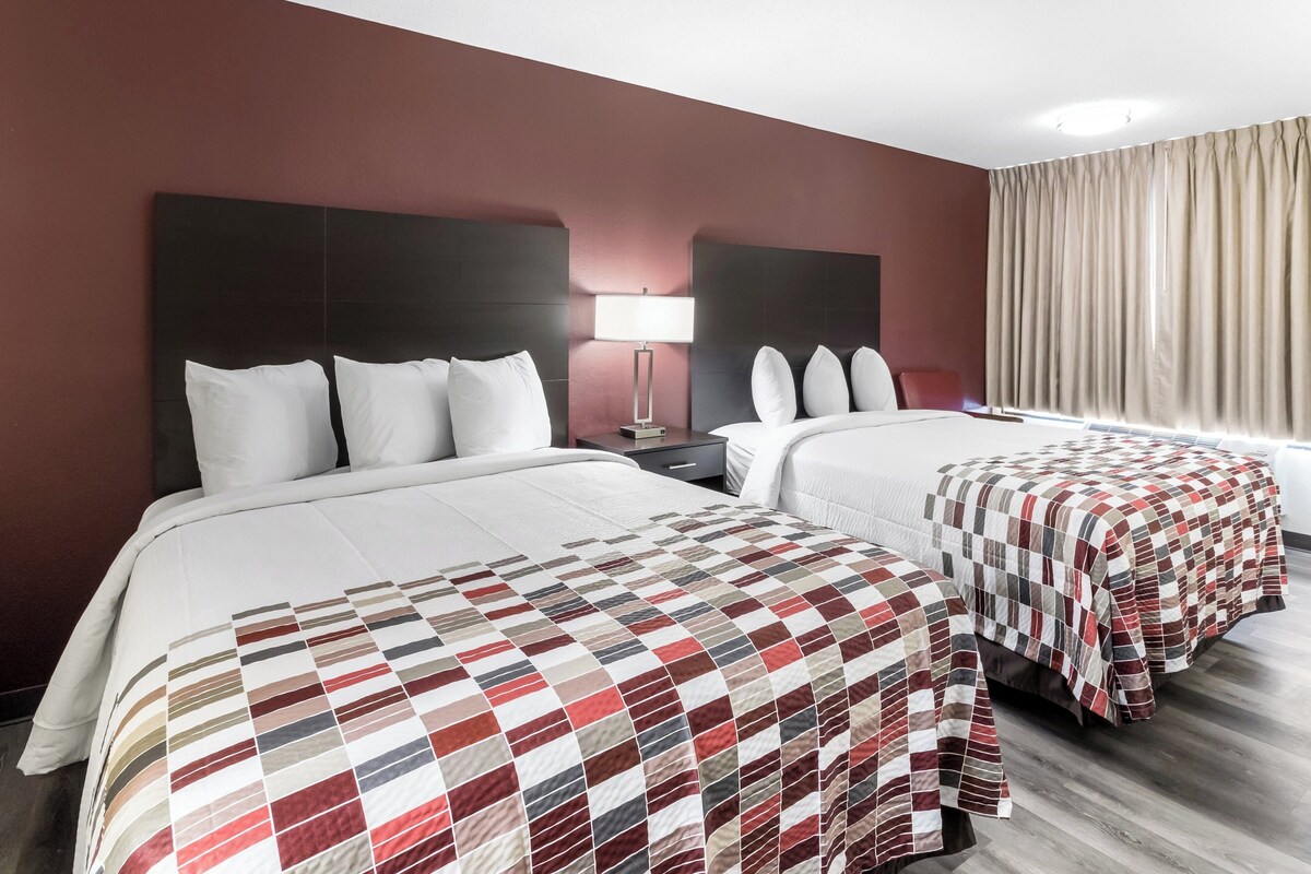 A hotel room with two beds, a nightstand, a lamp, and a window with curtains.