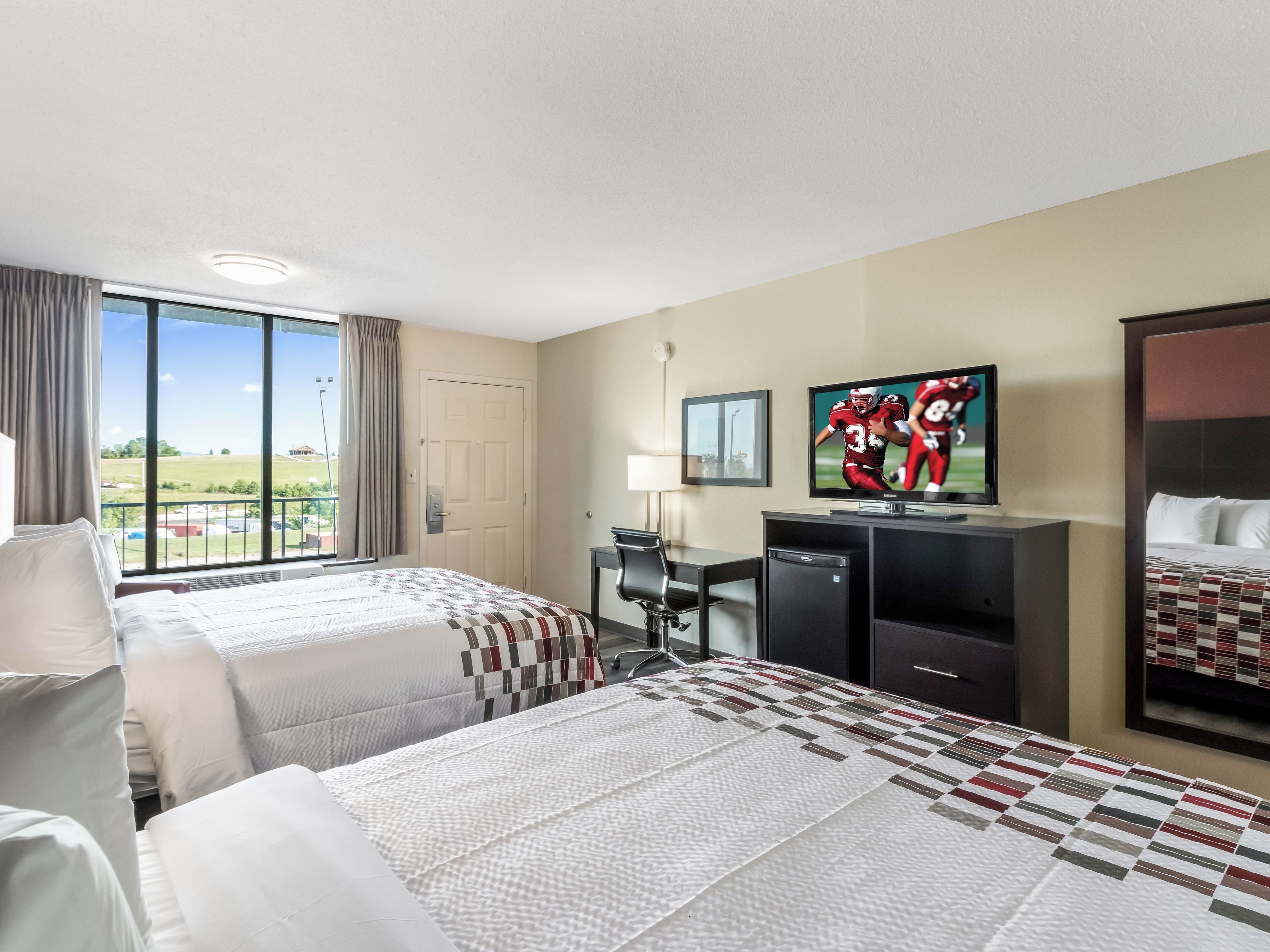 A hotel room with two beds, a desk, a television, and a view of the outdoors.