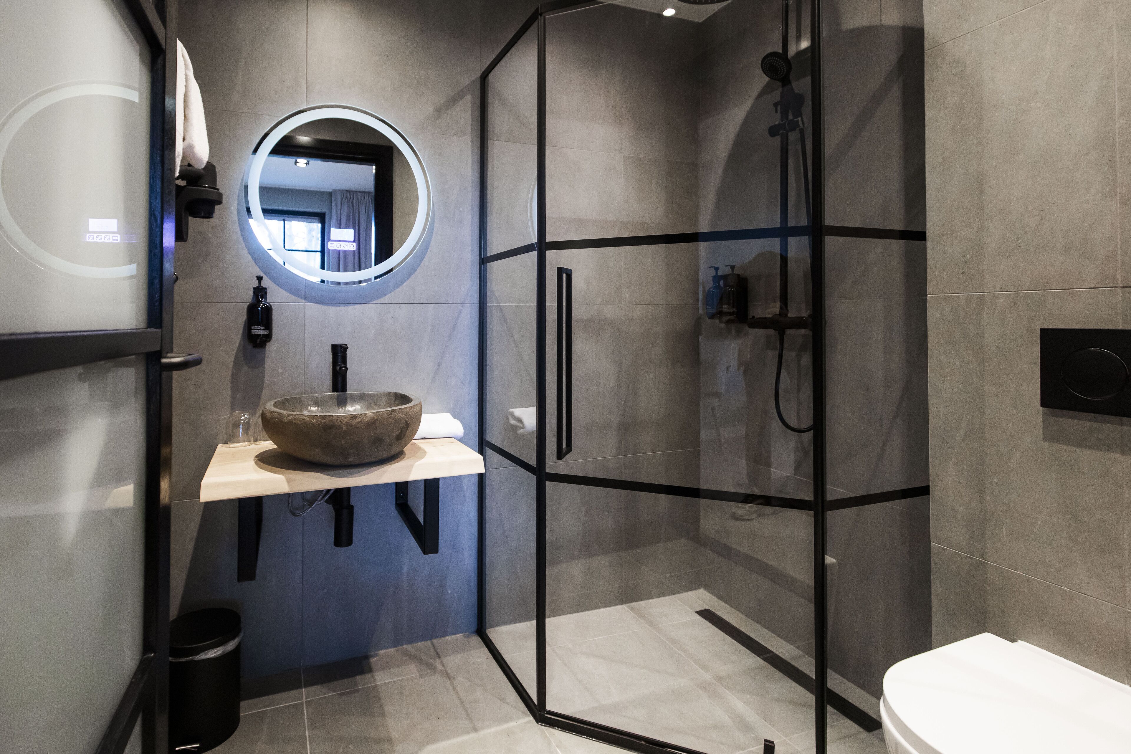 A modern bathroom with a glass-enclosed shower, a round mirror, a stone sink, and a toilet.