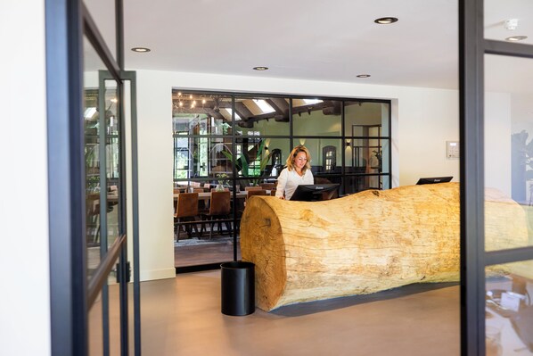 A modern interior with a large wooden sculpture, a reception desk, and a dining area visible through glass doors. - Veluwe Hotel de Beyaerd (Hulshorst)