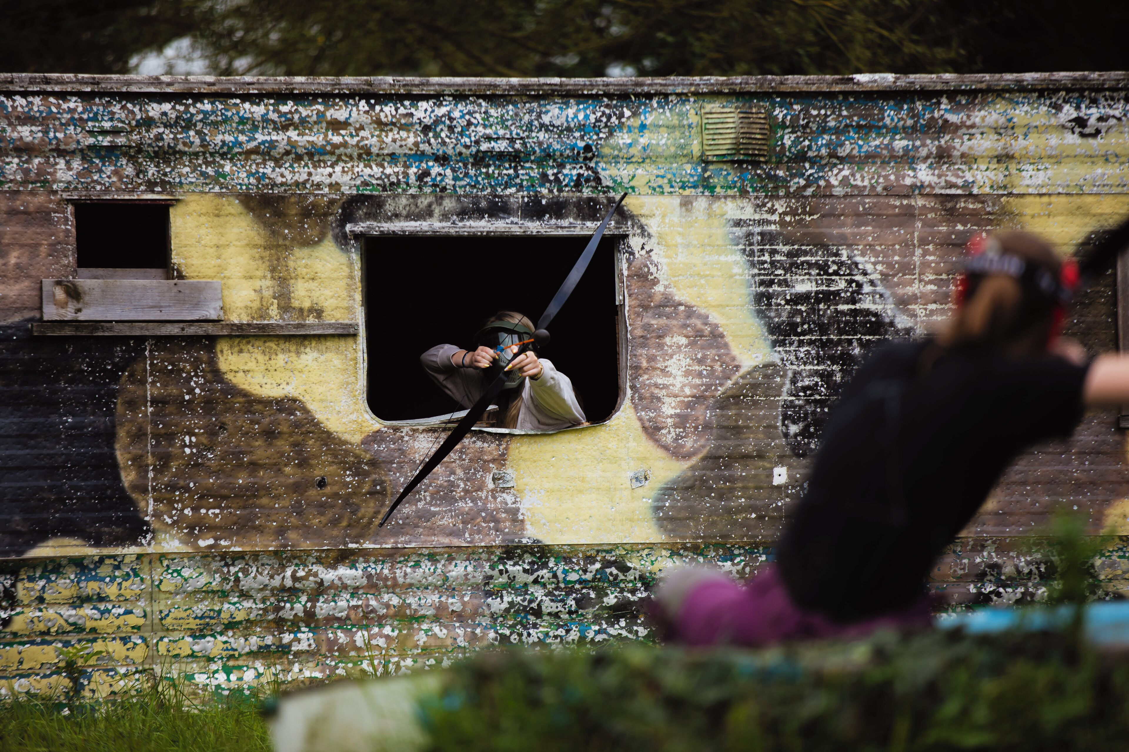 A person aiming a paintball gun through a window of a building with peeling paint and graffiti.